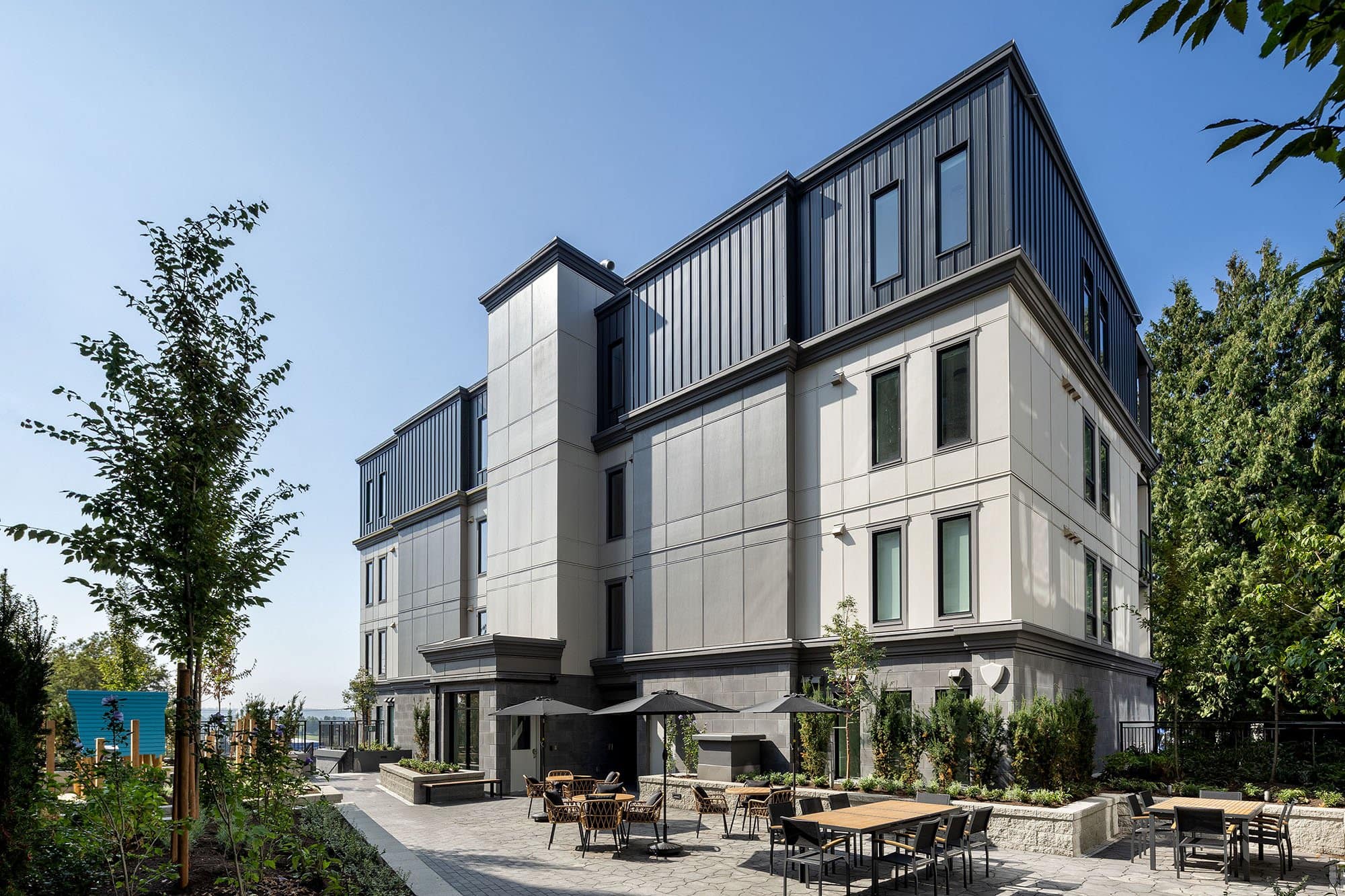 Outdoor courtyard amenity space at Bastille by Porte Homes located at 209 Lebleu Street in Coquitlam’s Maillardville neighbourhood featuring landscaped pathways, patio seating areas, outdoor dining tables, and modern building architecture surrounding the shared resident space.