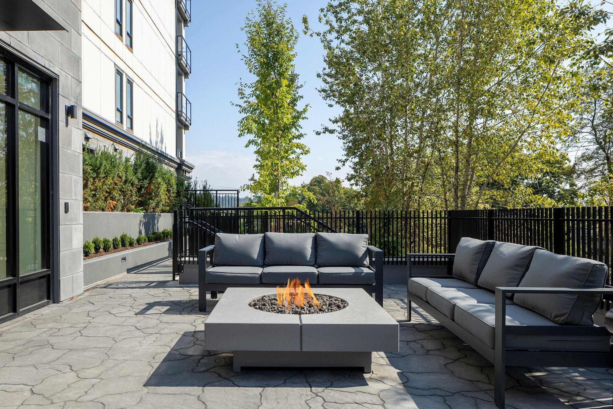Outdoor patio amenity at Bastille presale development by Porte Homes in Coquitlam’s Maillardville neighbourhood featuring modern outdoor sofas arranged around a built-in firepit table, stone paver patio flooring, landscaped trees, and a fenced seating area beside the residential building at 209 Lebleu Street.