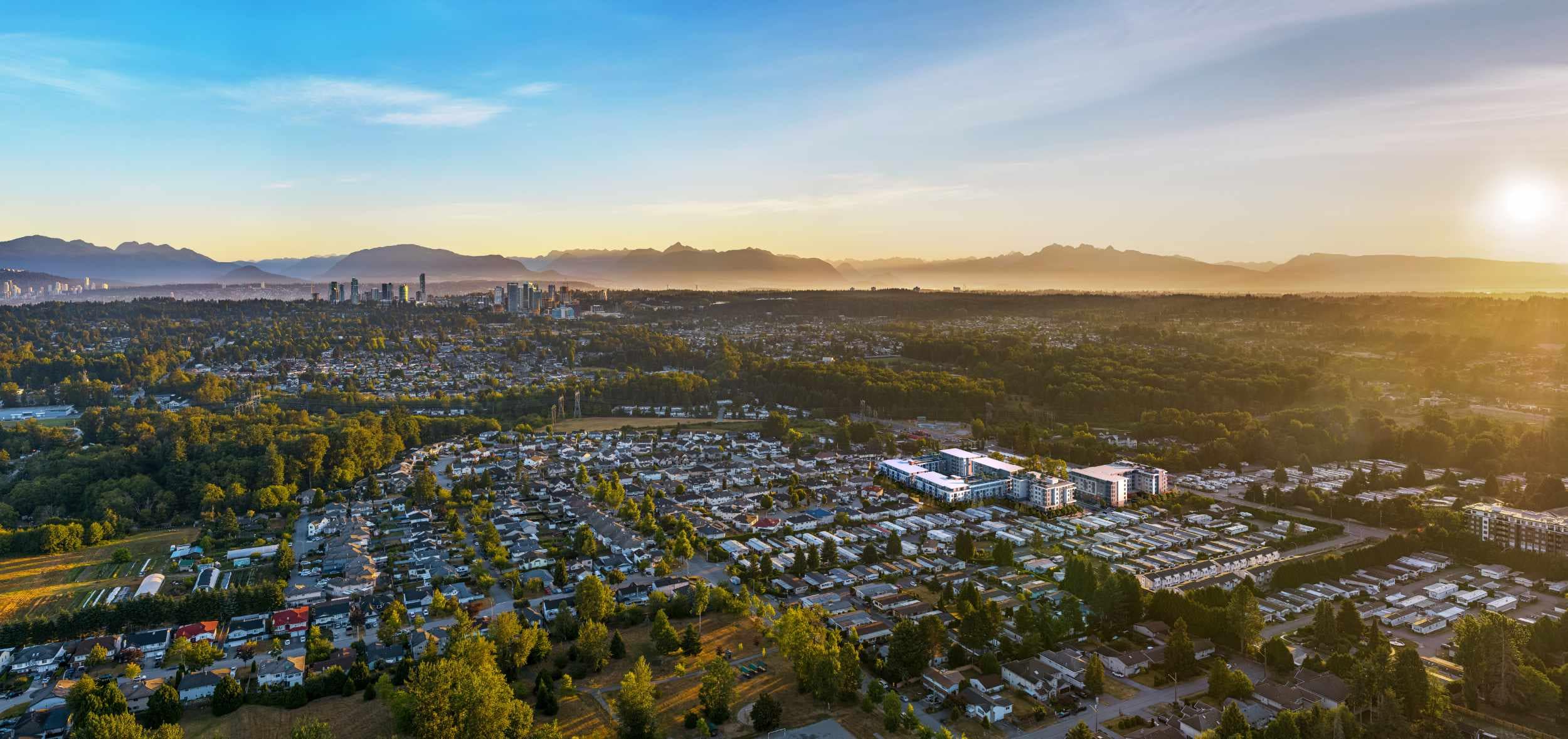 Aerial neighbourhood view near Spera presale development in Surreyโs Newton area by Peterson Group at 8293 King George Blvd, showing surrounding low-rise residential streets, tree-lined blocks, nearby parks and green space, distant Surrey skyline, and a mountain backdrop in warm sunset light, highlighting the developmentโs setting and connectivity.