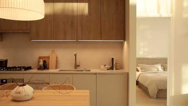 Video thumbnail showing a warm contemporary kitchen interior at Soenhause Coquitlam, a presale condominium development by Marcon located at 712 Como Lake Avenue in Coquitlam. The space features natural wood upper cabinetry, minimalist lower cabinets, integrated lighting, and a sleek kitchen sink and faucet. A dining table sits in the foreground while a bedroom is visible through an open doorway, illustrating the modern open-concept interior design planned for homes at Soenhause Coquitlam.