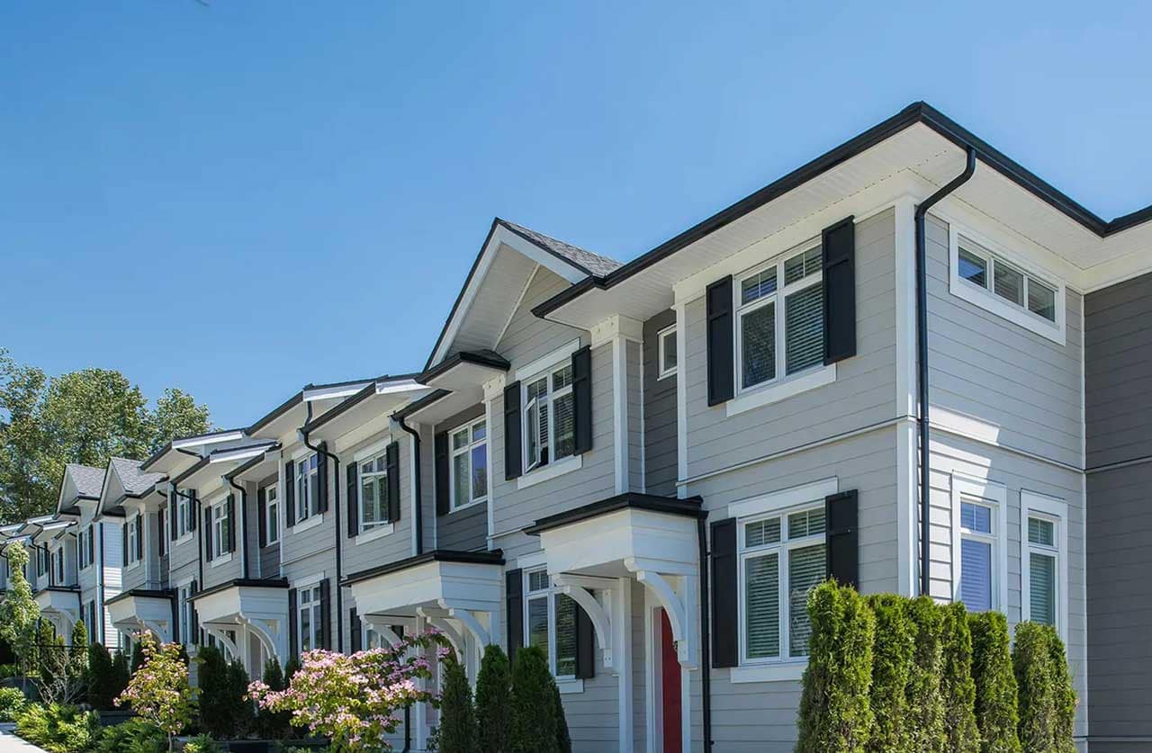 Exterior view of Chelsea Surrey, a presale townhome development by StreetSide Developments at 16325 21 Avenue in Surrey, featuring contemporary farmhouse-inspired homes with white and grey siding, black shutters, landscaped front gardens, and tree-lined walkways.