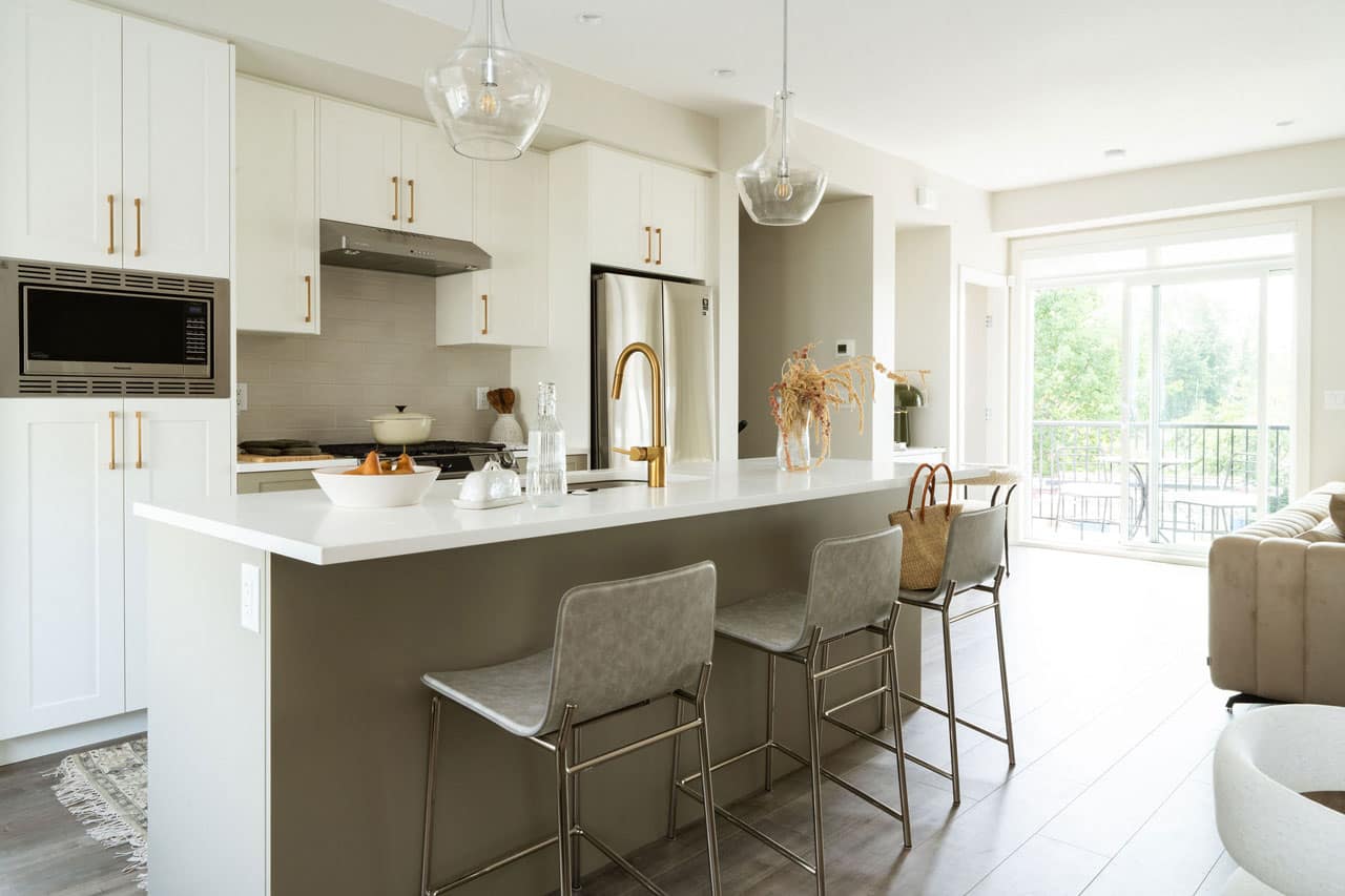 Open-concept kitchen at Fox & Aikins in South Surrey by Park Ridge Homes, 1871 165A Street, featuring a large quartz island with bar seating, gold kitchen faucet, stainless steel appliances, white shaker cabinetry, pendant lighting, and seamless flow to the living and balcony area.