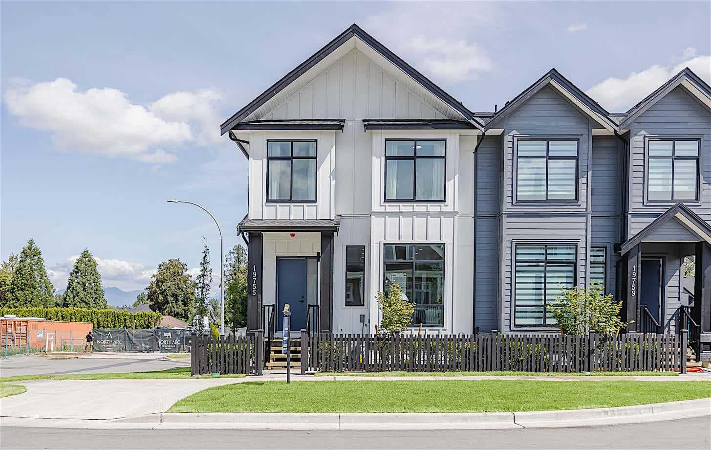 Front exterior of townhomes at Latimer Landing Langley by Archstone, located at 7760 196 Street. The homes feature modern architecture, pitched rooflines, large windows, fenced front yards, landscaped green space, and attractive curb appeal in a family-oriented community.