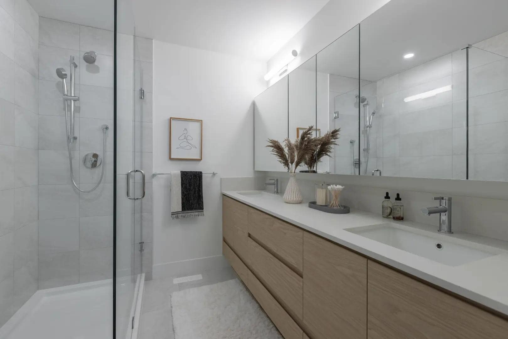 Modern bathroom interior at Chelsea at The Boroughs presale townhomes in Surreyโs Sunnyside Heights neighbourhood, developed by Northwest Development at 16325 21 Avenue, featuring a long double vanity with light wood cabinetry, quartz countertop, large wall mirror, contemporary lighting, and a glass walk-in shower with chrome fixtures.