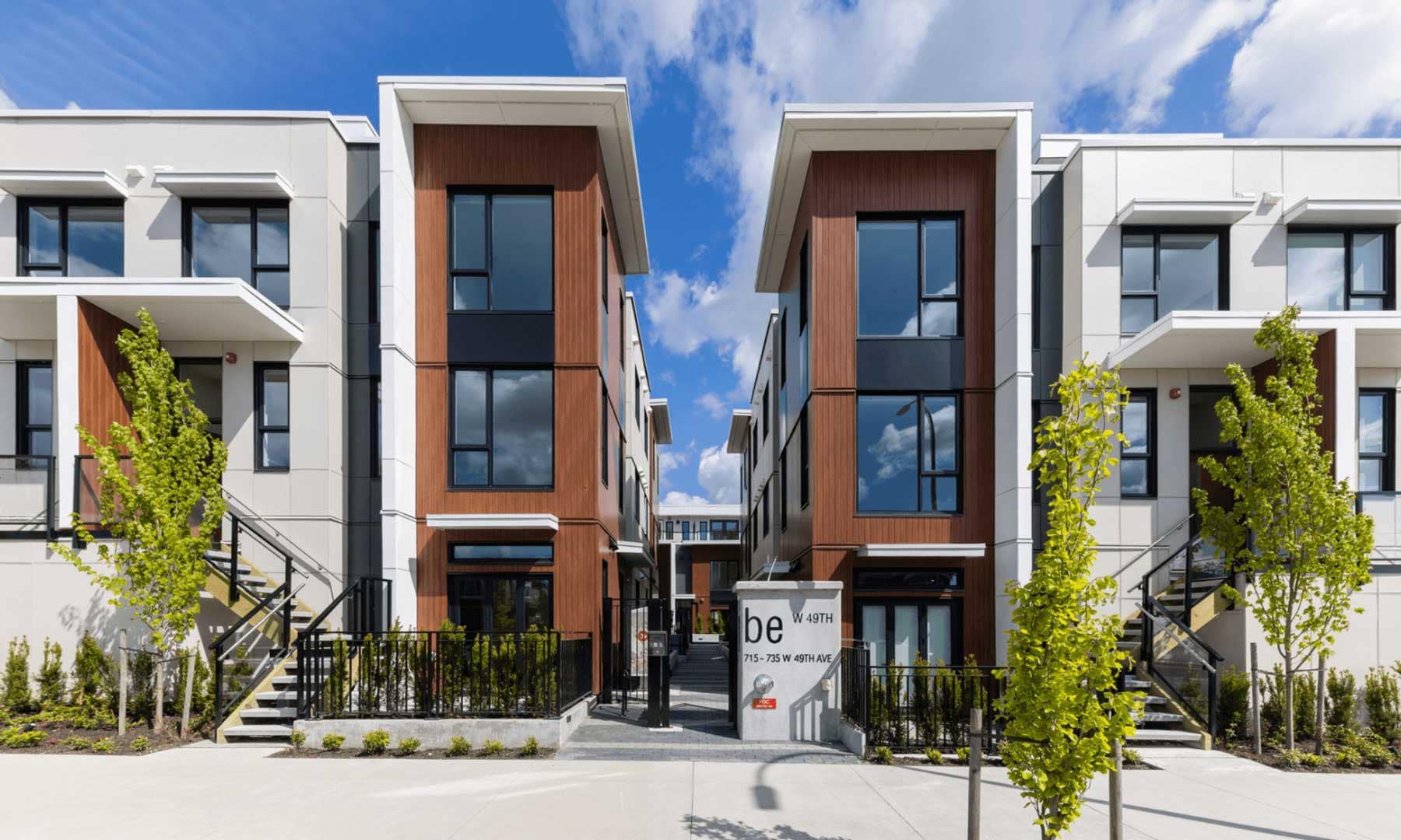 Exterior view of Be W 49th Vancouver by Modella Developments at 715 West 49th Avenue in Oakridge, featuring modern three-storey townhomes with wood-accented faรงades, large black-framed windows, landscaped walkways, and contemporary West Side architecture.