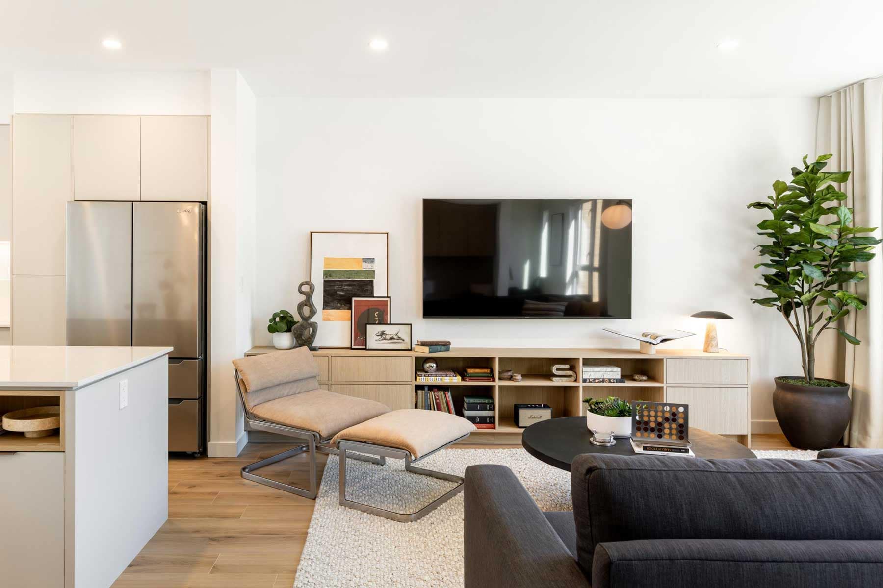 Open-concept living space in a Towns at Burke Coquitlam townhome showing a lounge chair, wood media console, stainless fridge, and a fiddle-leaf fig plant.