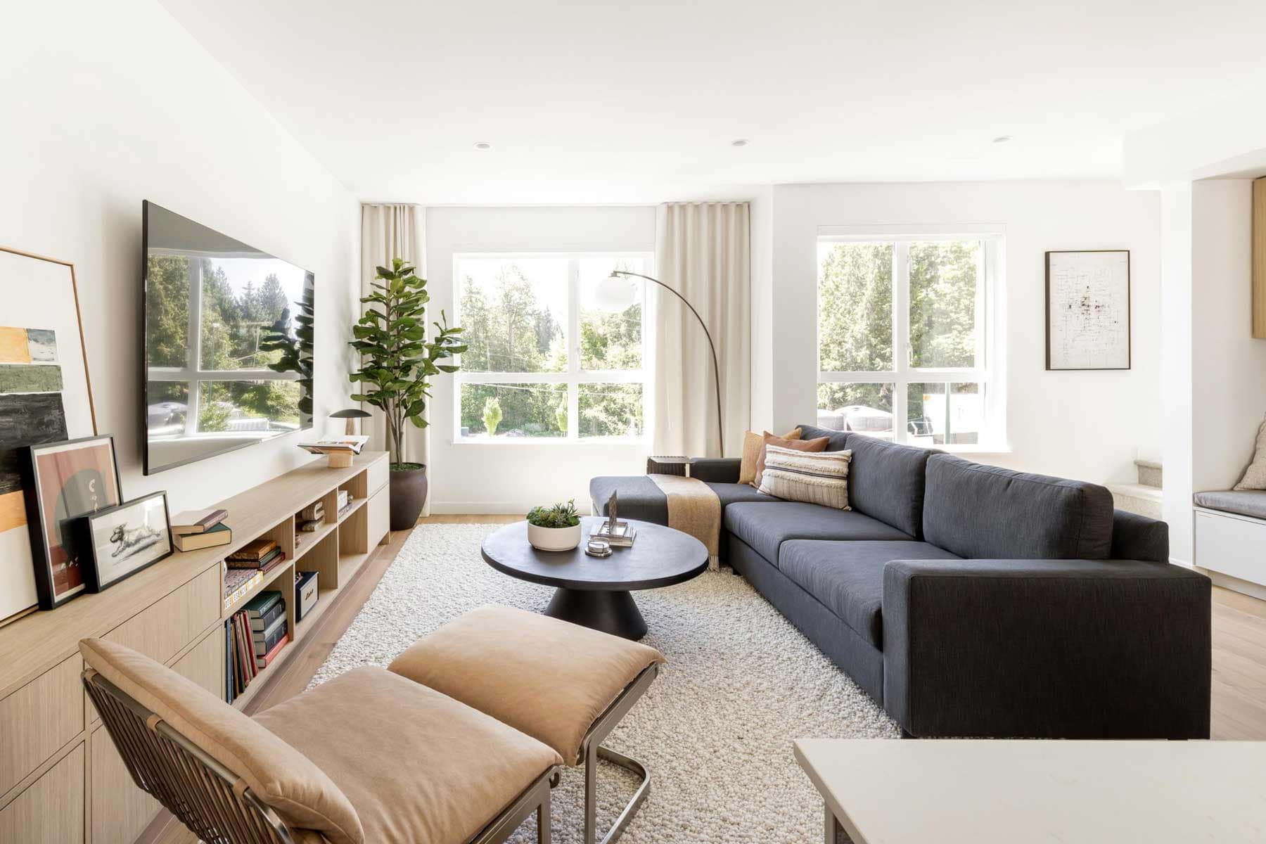 QuiBright living room in a Towns at Burke Coquitlam townhome with a dark grey sectional, wall-mounted TV, wood console, and large windows facing trees.