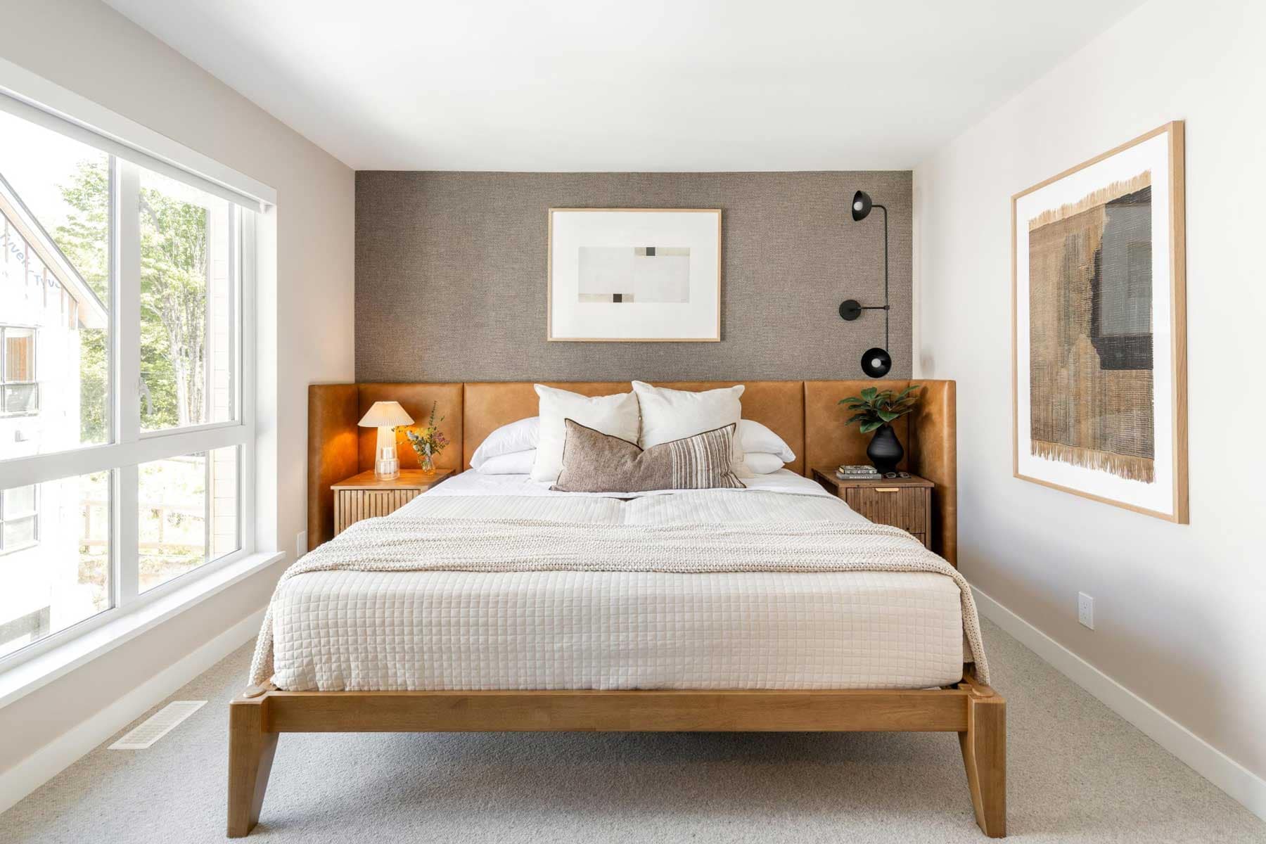 Primary bedroom in a Towns at Burke Coquitlam townhome with a caramel leather headboard, wood bed frame, wall sconce lighting, and a large window with treed views.