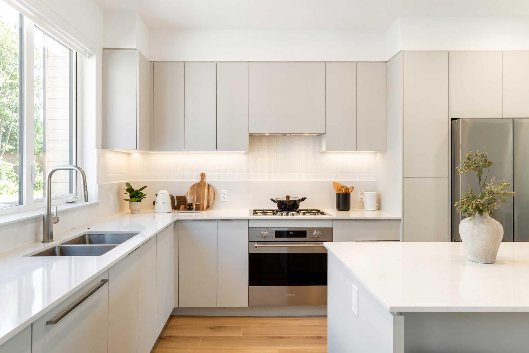 Modern kitchen in a Towns at Burke Coquitlam townhome with light grey cabinetry, a gas range, stainless fridge, and quartz countertops lit by a large window.