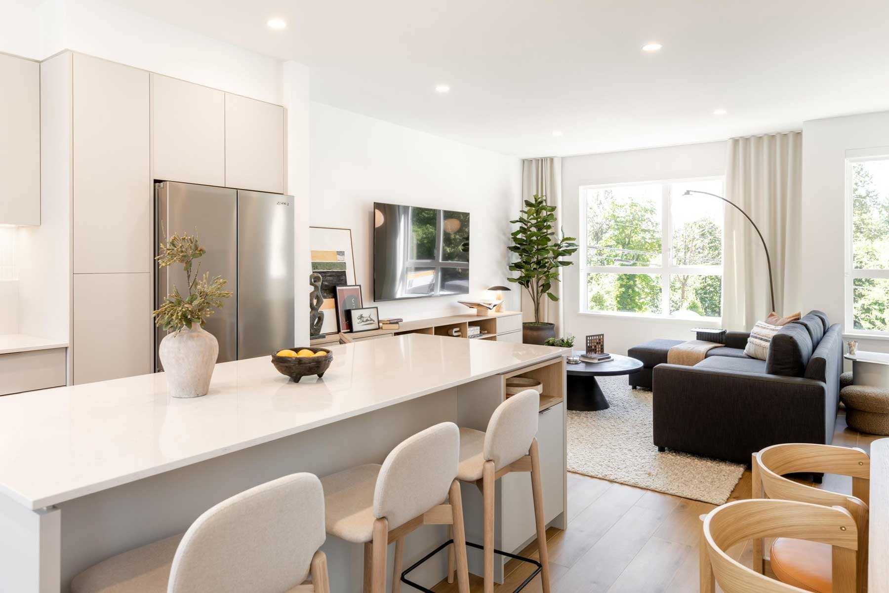 Open-concept main floor in a Towns at Burke Coquitlam townhome with a large kitchen island, bar stool seating, stainless fridge, and a living room with sectional and floor-to-ceiling windows.