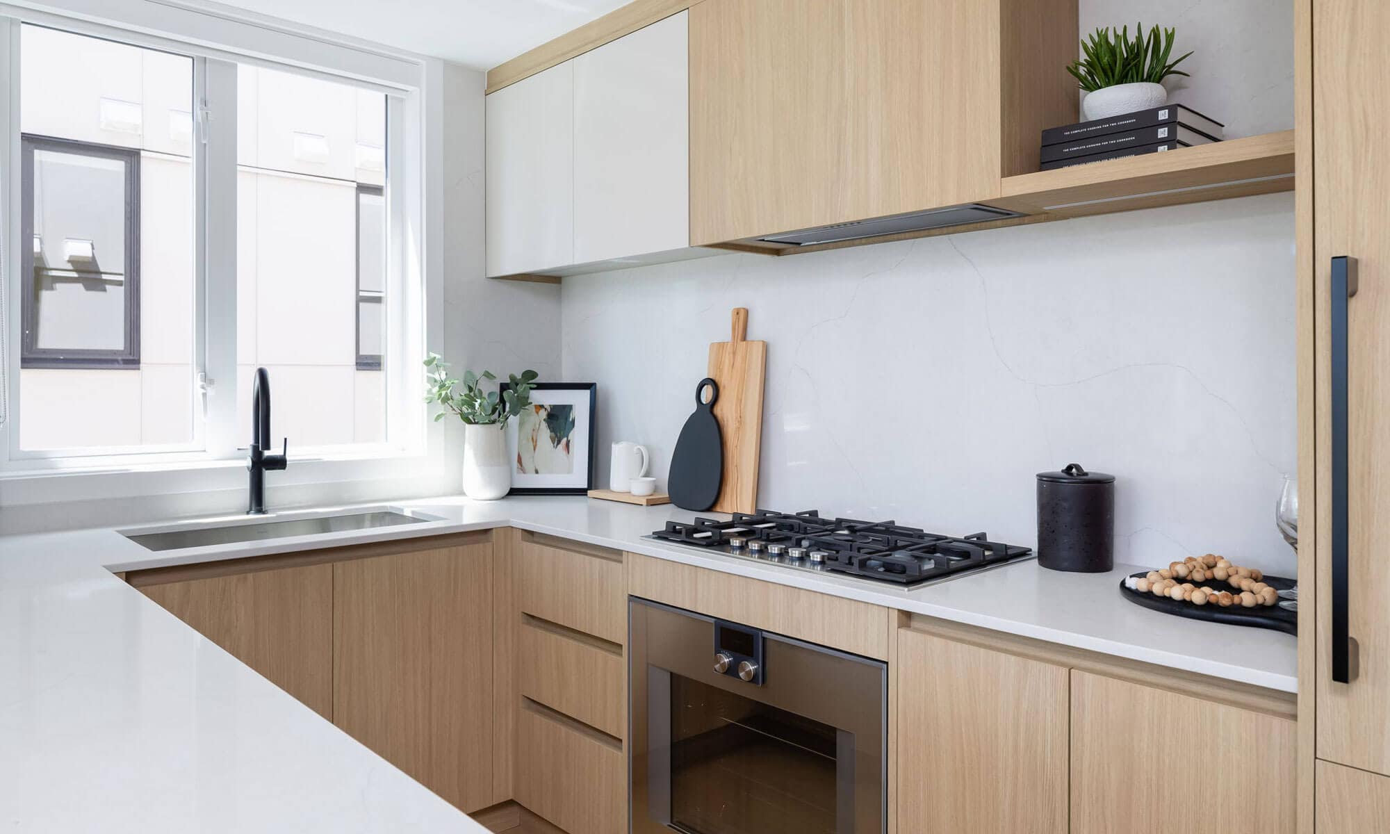 Bright contemporary kitchen interior at Be W 49th Vancouver by Modella Developments at 715 West 49th Avenue in Oakridge, featuring light oak cabinetry, white stone countertops, integrated appliances, black fixtures, and large windows bringing natural light into the space.