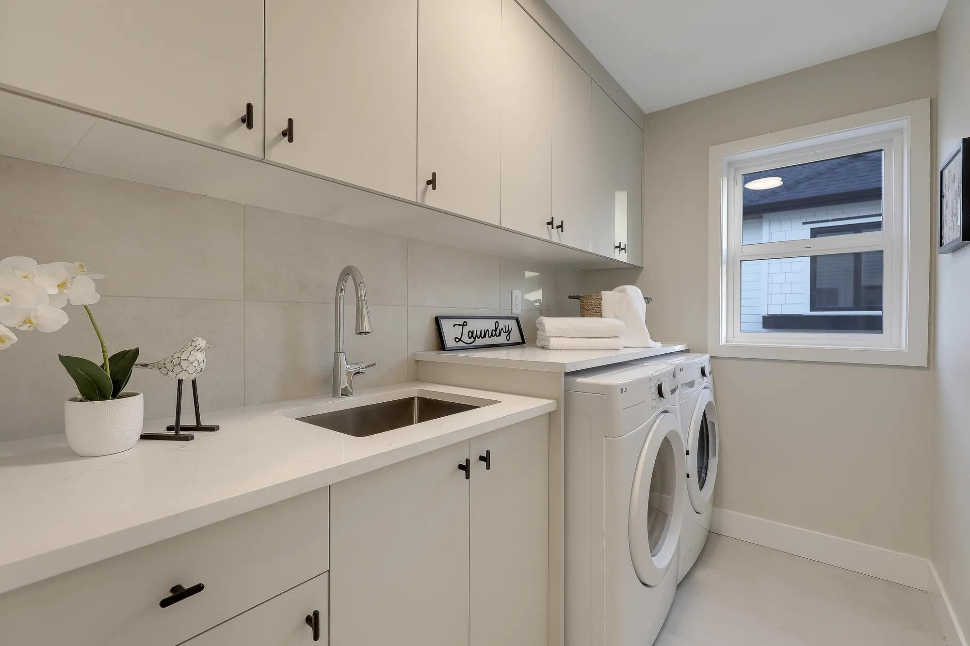 Dedicated laundry room at Latimer Landing Langley by Archstone featuring side-by-side washer and dryer, utility sink, quartz countertop, upper storage cabinets, modern fixtures, and a bright window providing natural light.