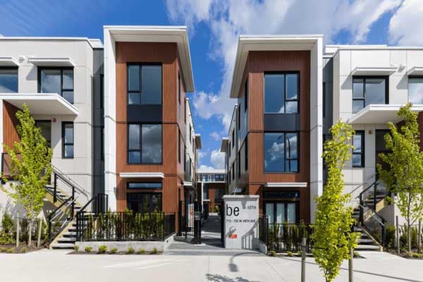 Front entrance and streetscape view of Be W 49th Vancouver in Oakridge by Modella Developments, showcasing modern townhouse architecture, landscaped frontage, private walk-up entries, and the development signage at 715 West 49th Avenue.